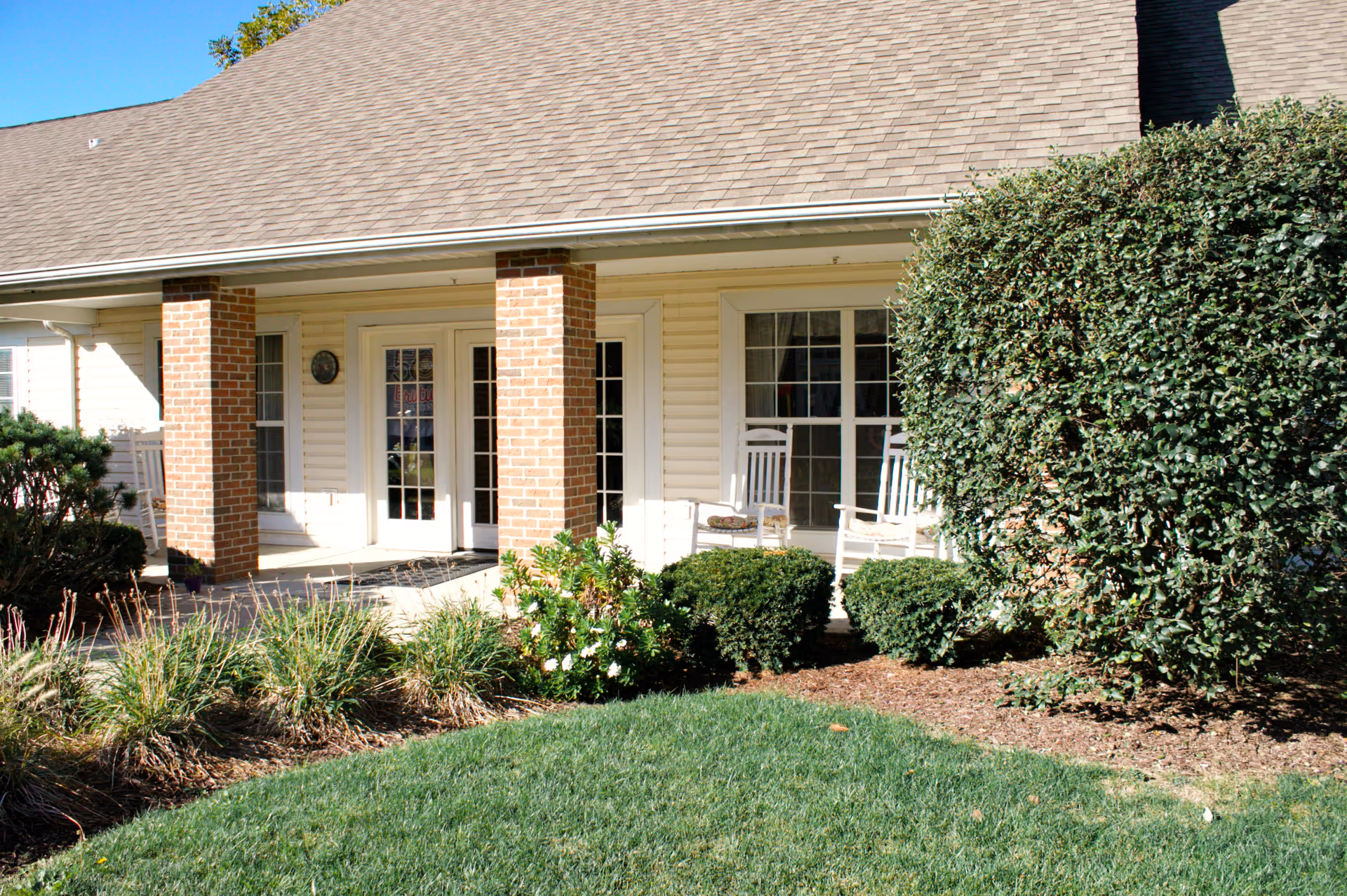 Exterior view of a senior living facility porch area with two white rocking chairs, brick columns, large windows, and well-maintained bushes and grass in front.