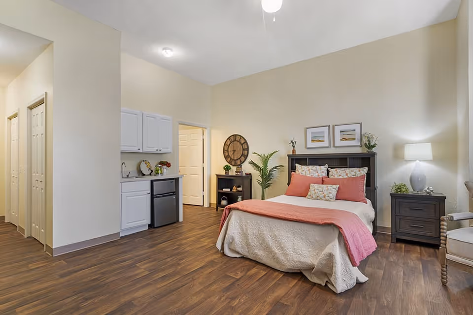 A well-lit bedroom in a senior living facility featuring a bed with white and coral bedding, two framed pictures above the headboard, a nightstand with a lamp and plant, a small kitchenette with white cabinets and a mini fridge, a wooden floor, and a hallway with closed doors.
