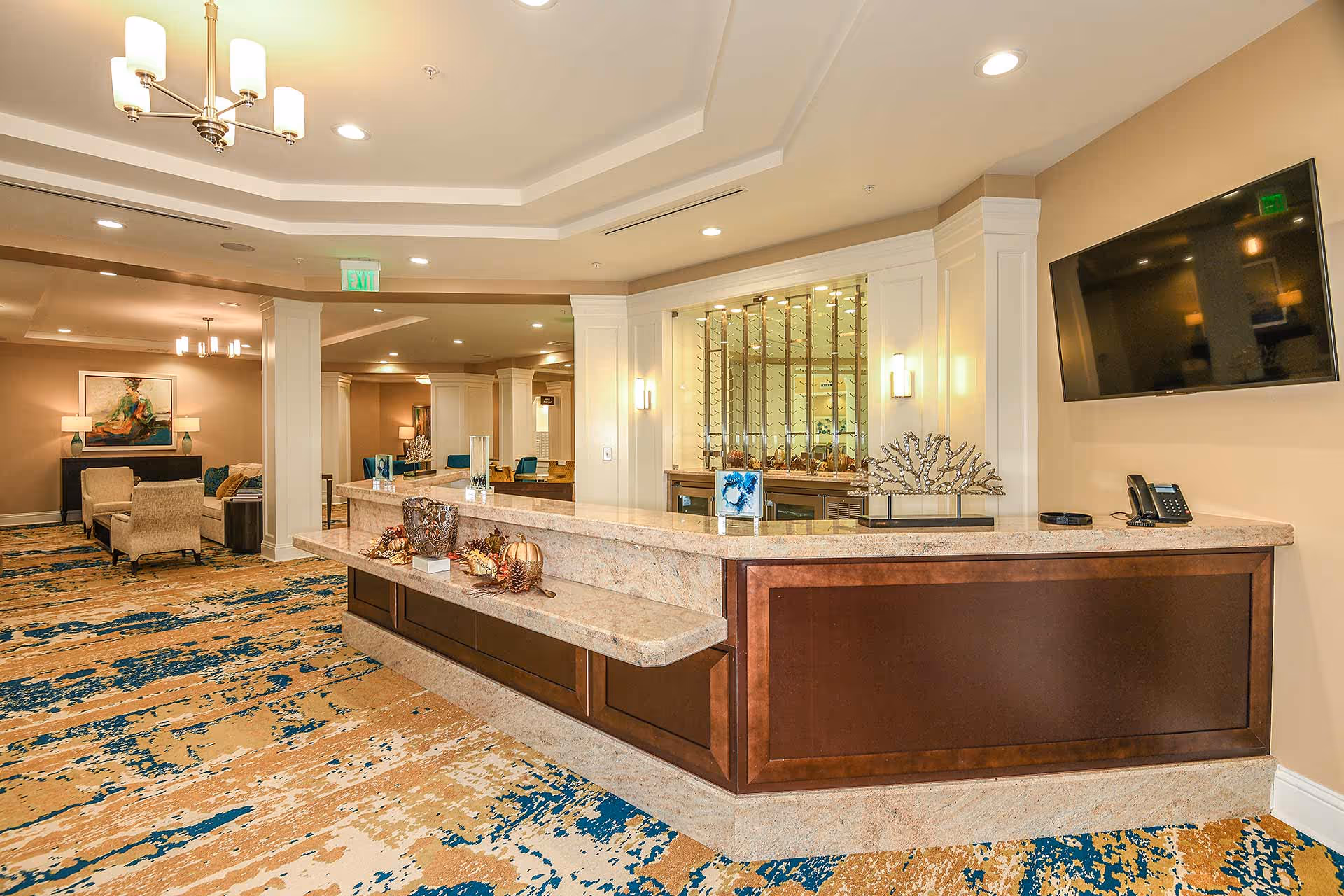 Reception area in a senior living facility with a large marble countertop desk decorated with ornamental items, a mounted flat-screen TV on the wall, and a seating area with chairs and artwork in the background. The space features warm lighting and a patterned carpet.
