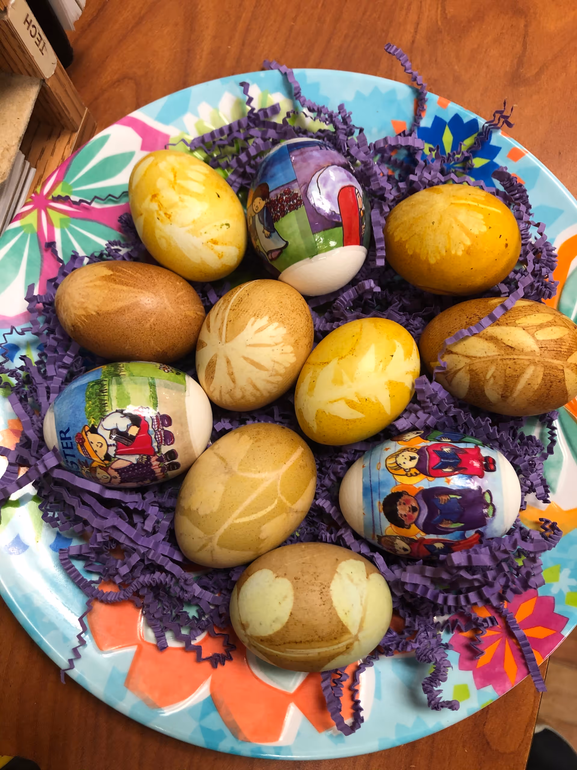 A colorful plate of decorated and naturally dyed Easter eggs nestled in purple paper shreds on a wooden table.