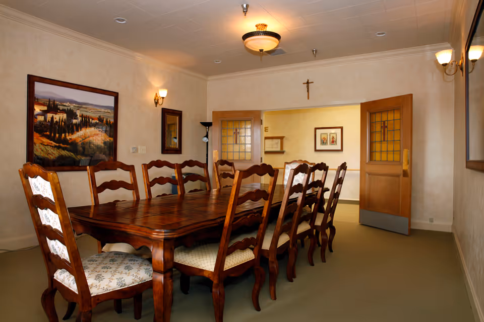 A dining room with a long wooden table surrounded by eight wooden chairs with patterned cushions. The room has beige walls, a ceiling light fixture, wall sconces, a large framed landscape painting, and a crucifix above double wooden doors with stained glass panels.