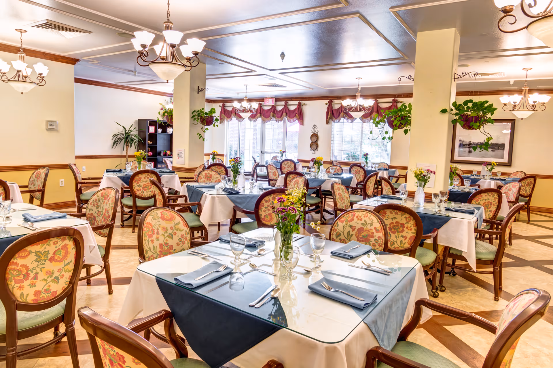 Bright, elegant dining room with multiple tables set with place settings, floral-upholstered chairs, hanging plants, and chandeliers.