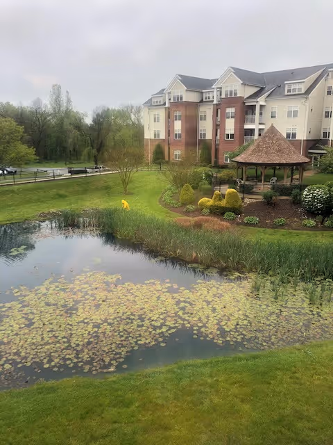 A senior living facility building with multiple floors and a mix of brick and light-colored siding. In front of the building is a landscaped garden area with trimmed bushes and a gazebo. A pond with lily pads is in the foreground, surrounded by green grass and trees in the background under an overcast sky.