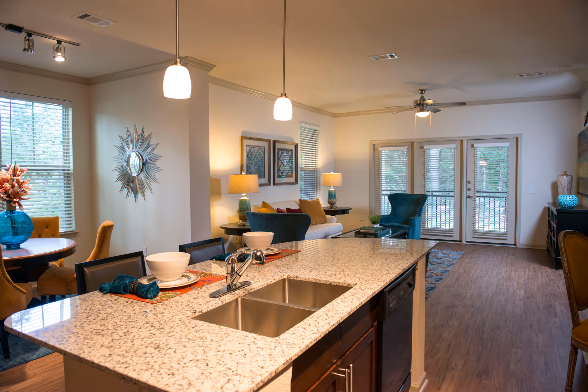 View of a modern open-concept living space in a senior living facility, featuring a granite kitchen island with a double sink and place settings, a dining area with a round table and chairs, a living room with teal and mustard accent chairs, a white sofa, lamps, wall art, and glass doors leading to an outdoor area.