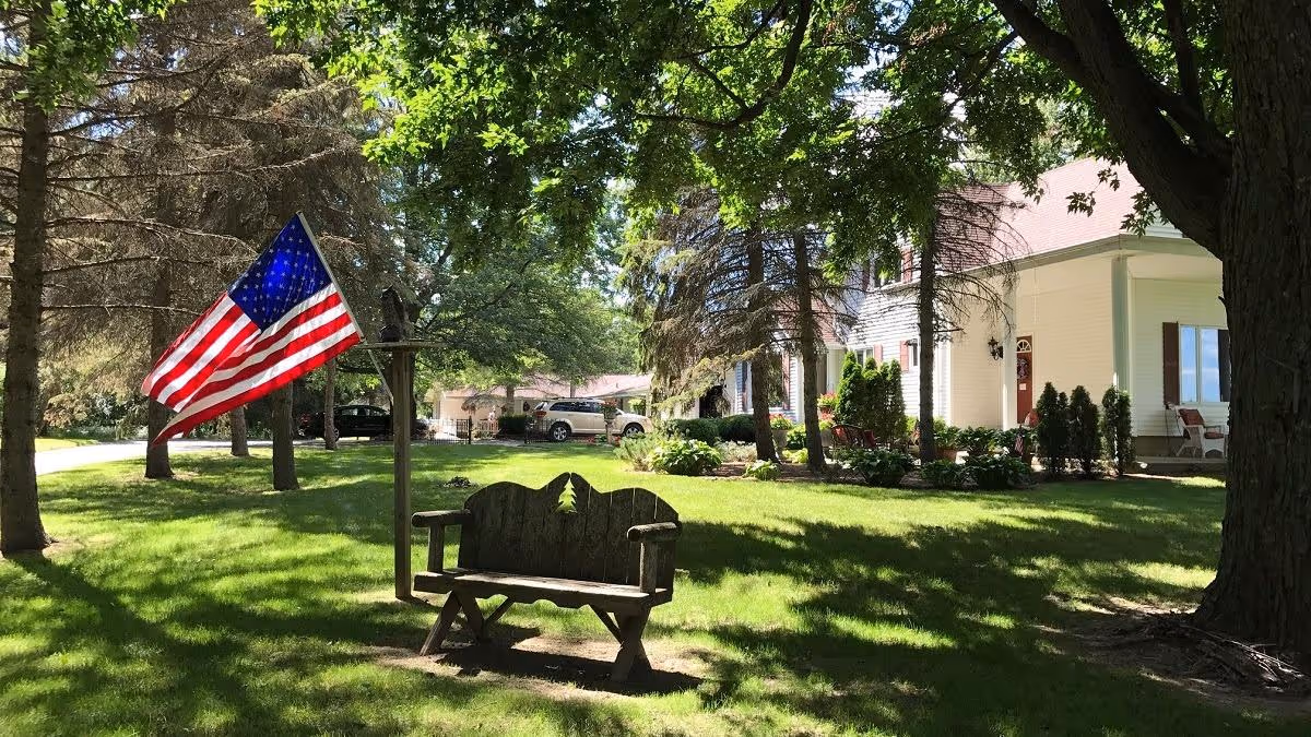 A sunny outdoor scene at Pine Haven Senior Assisted Living featuring a wooden bench with a decorative backrest, an American flag on a pole, lush green grass, tall trees providing shade, and a white building with a porch in the background.
