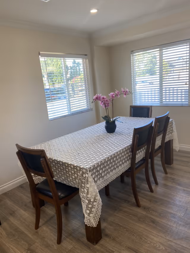A dining room with a rectangular wooden table covered by a white lace tablecloth. Four wooden chairs with black cushions surround the table. A small black vase with pink orchids is placed in the center of the table. Two windows with white blinds allow natural light into the room, which has light-colored walls and wooden flooring.