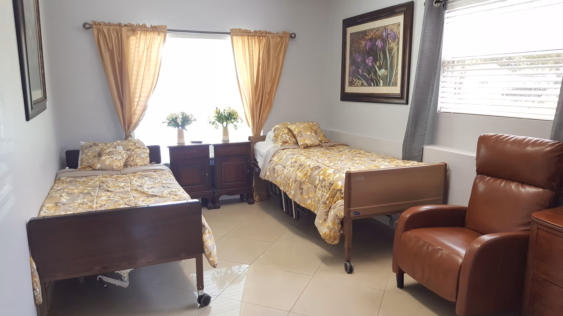 A bright room with two single beds covered in matching yellow floral bedspreads, a brown leather armchair, two wooden nightstands with flower vases, beige tiled floor, and windows with curtains allowing natural light to enter.