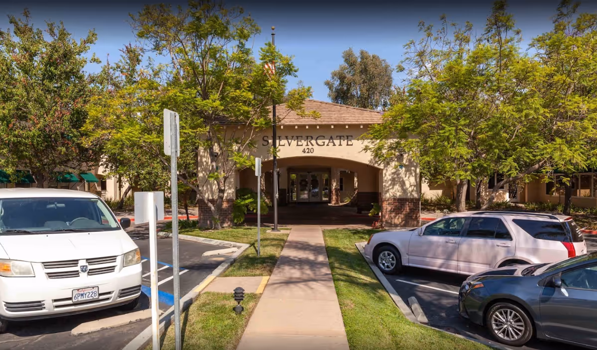 Front entrance of Silvergate Fallbrook Retirement, with trees, parked cars, and walkway.