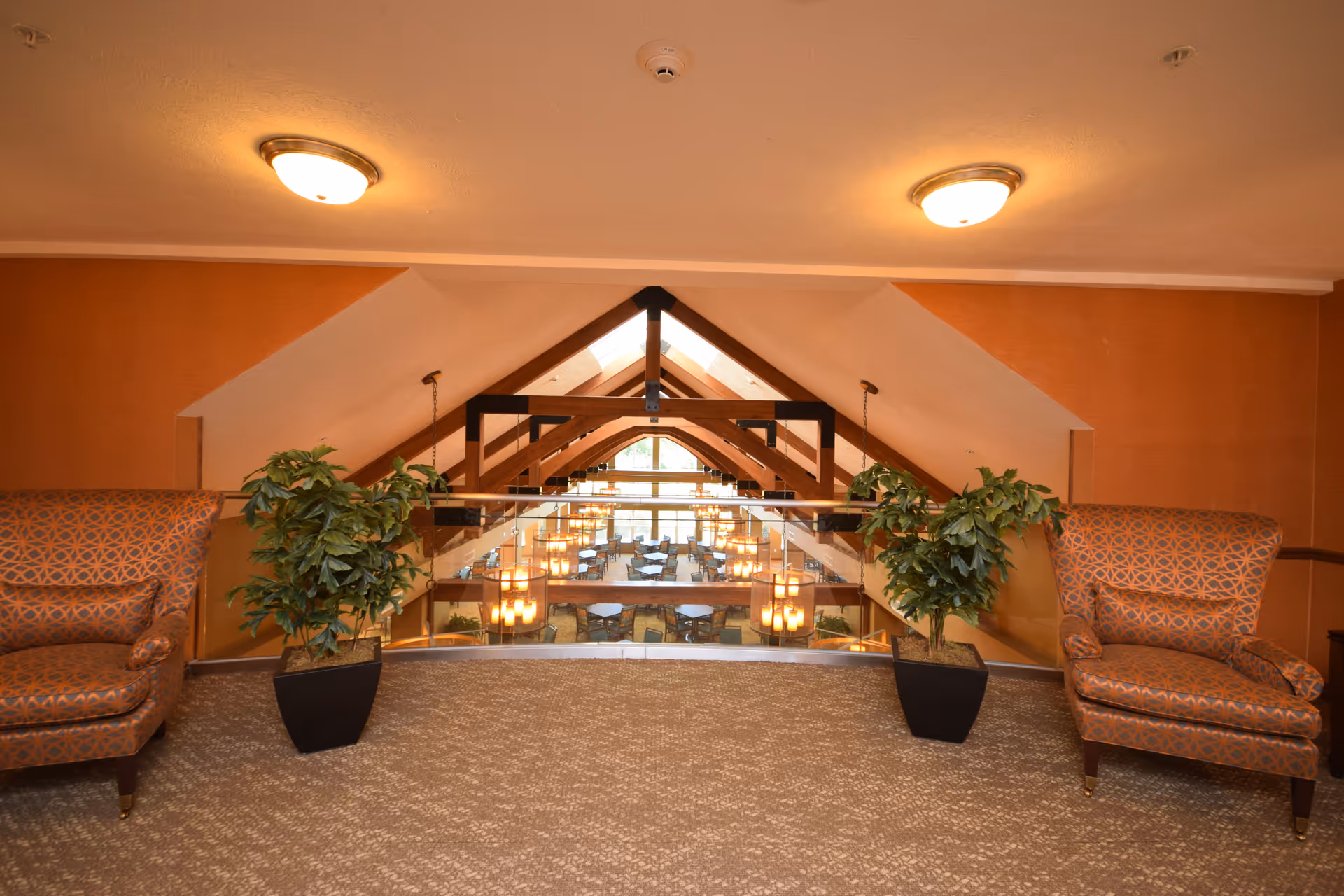 Interior view of a senior living facility showing a carpeted seating area with two patterned armchairs and two potted plants on either side. The area overlooks a dining room below with multiple tables and chairs, visible through a glass railing. The ceiling features wooden beams and hanging lights.