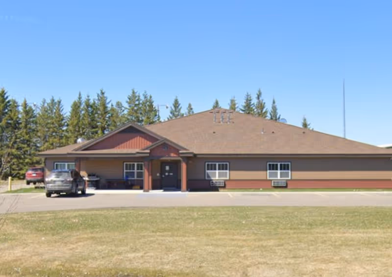 Single-story building with a brown roof and brown siding, featuring a small covered entrance with two pillars. There are two cars parked in front of the building, and a row of evergreen trees in the background under a clear blue sky.