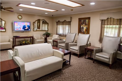 A cozy living room with beige walls and carpeted floor featuring a white loveseat, four beige armchairs, wooden side tables, a wall-mounted flat screen TV showing a game show, decorative mirrors, framed artwork, and windows with curtains.