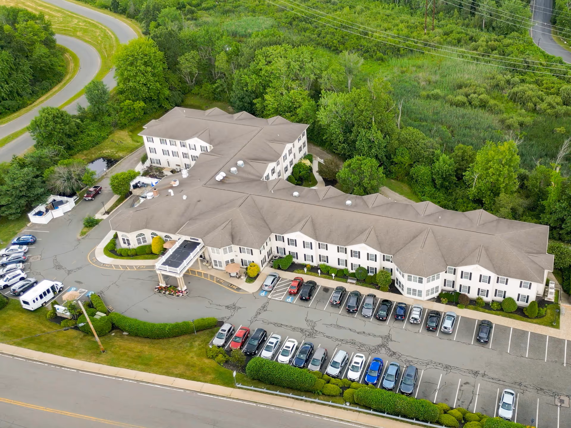 Aerial view of Benchmark Senior Living at Waltham Crossings, showing a large two-story building surrounded by greenery and trees. The building has a beige roof and white exterior walls with multiple windows. There is a parking lot in front of the building with several cars parked, and a curved road leading to the entrance. The surrounding area includes a road, grassy areas, and dense vegetation.