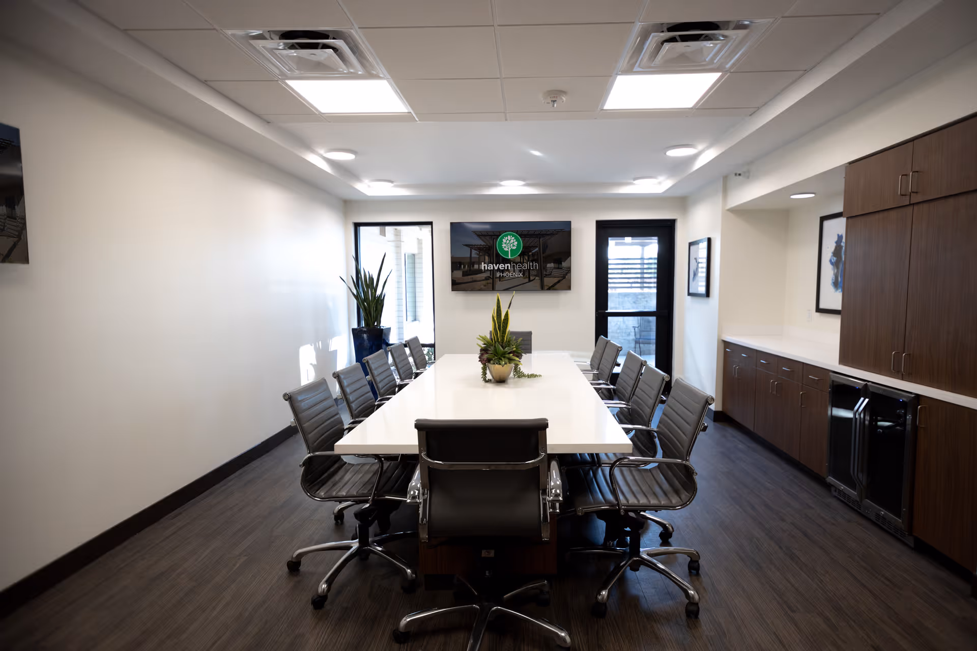 A modern conference room with a long white table surrounded by black office chairs on wheels. There are two large windows and a glass door at the far end of the room. A flat-screen TV mounted on the wall displays the Haven Health Phoenix logo. The room has wooden cabinetry along one wall, a potted plant on the table, and recessed ceiling lights.