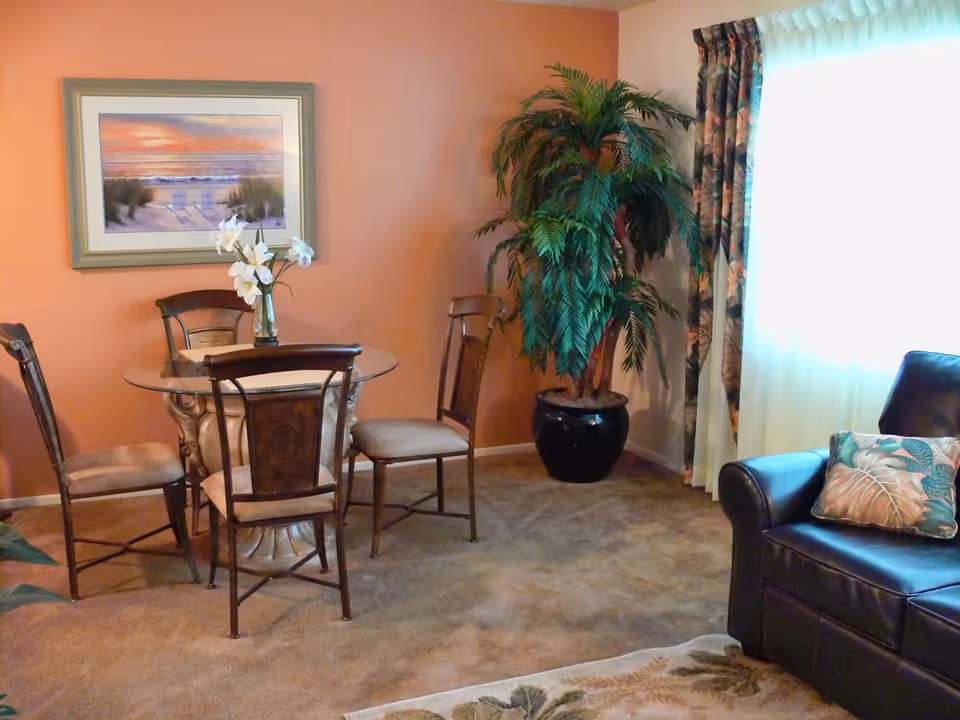 A cozy living area with a round glass-top table surrounded by four wooden chairs with cushioned seats. A vase with white flowers is placed on the table. To the right, there is a large potted plant next to a window with floral curtains and sheer white drapes. A dark leather armchair with two decorative pillows is partially visible in the foreground. The walls are painted a warm peach color, and a framed beach sunset painting hangs on the wall above the table.