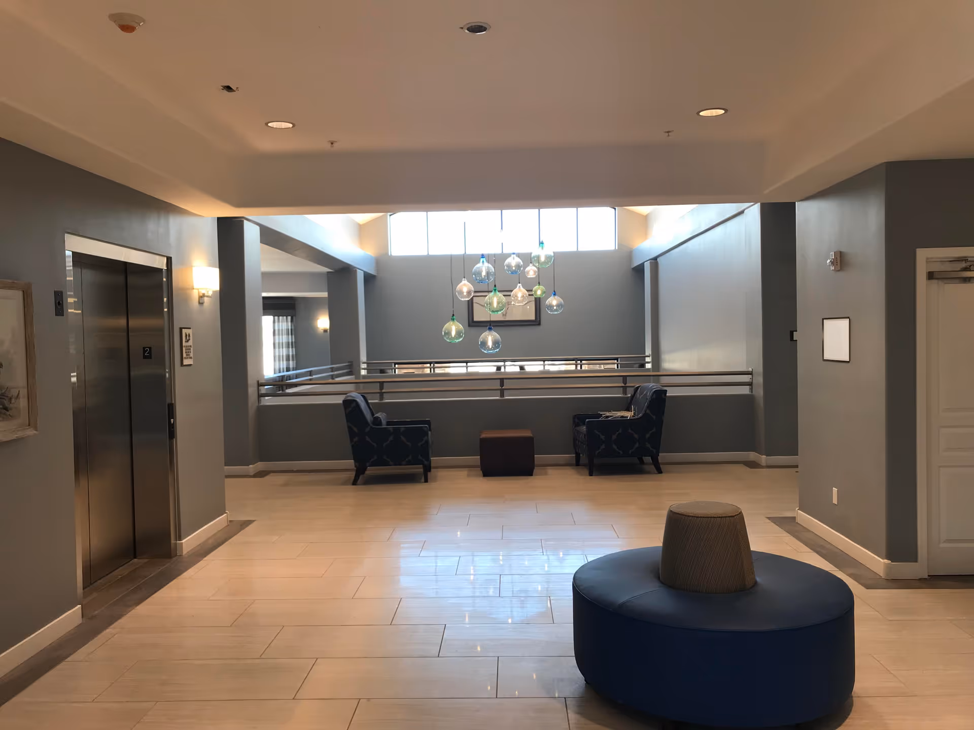 Interior view of a senior living facility hallway with an elevator on the left, two armchairs and a small table near a railing, and a round blue cushioned bench in the foreground. The walls are painted gray and there are hanging decorative glass light fixtures near a window letting in natural light.