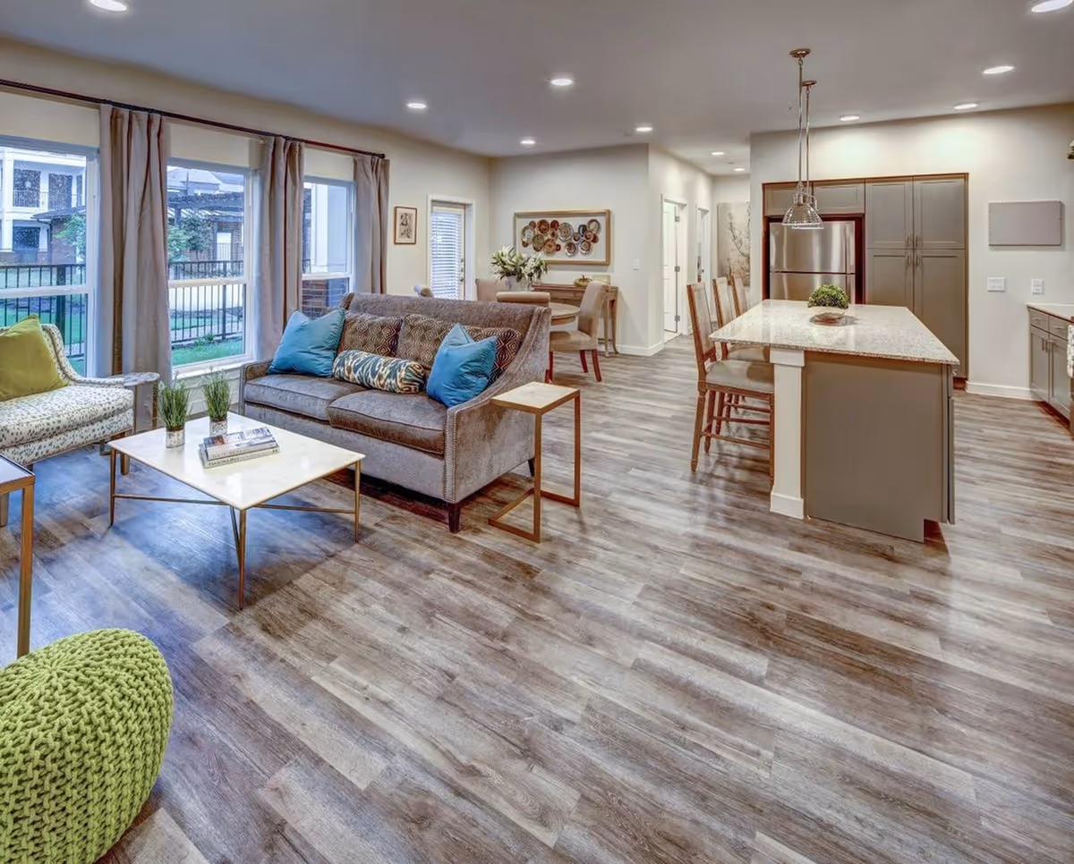 A modern open-concept living space featuring a living room with a gray sofa adorned with blue and patterned pillows, a white marble coffee table with small plants and books, a patterned armchair, and a green knitted pouf. The space extends into a kitchen area with a large island, bar stools, stainless steel refrigerator, and pendant lighting. Large windows with beige curtains allow natural light to fill the room, and a small dining area with a round table and chairs is visible in the background.