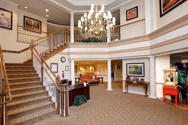 Two-story senior living lobby with a grand chandelier, sweeping staircase, reception desk, and seating area.