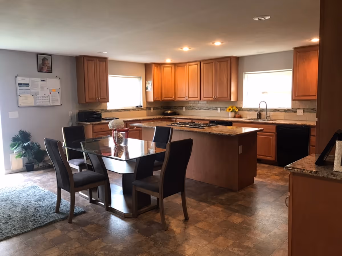 A spacious kitchen and dining area with wooden cabinets, granite countertops, and a central island with a built-in stove. The dining area features a glass-top table with six dark cushioned chairs. Two windows provide natural light, and there is a small plant and a bulletin board on the wall.