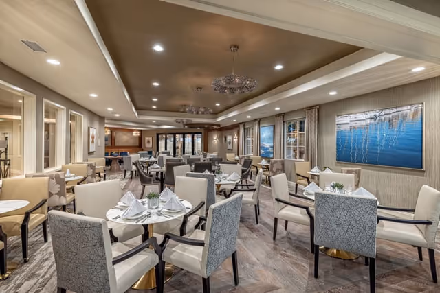 A spacious and elegant dining room with multiple round tables set with white napkins and glassware. The room features comfortable upholstered chairs, modern chandeliers hanging from a recessed ceiling, large windows with curtains, and artwork on the walls. The flooring is wood-patterned, and the overall ambiance is warm and inviting.