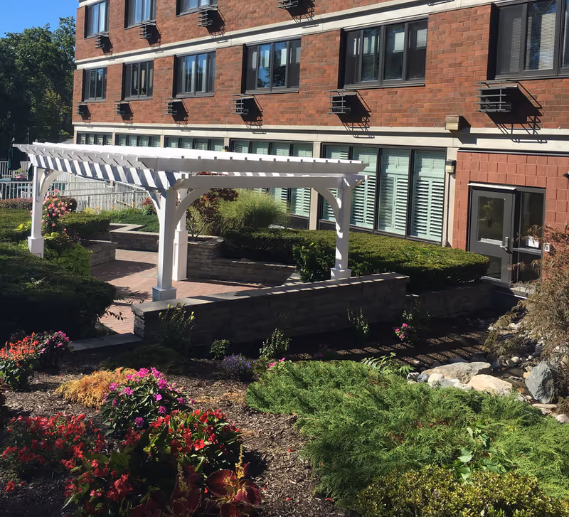 Outdoor garden area at Immanuel House featuring a white pergola over a brick-paved pathway, surrounded by various green shrubs, colorful flowers, and a stone retaining wall. The background shows a multi-story brick building with multiple windows and a door.