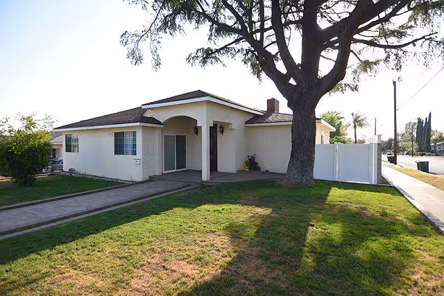 Single-story residential building with a covered entrance, large tree in front yard, and a paved walkway leading to the door. The building is surrounded by a grassy lawn and a white fence on the right side.