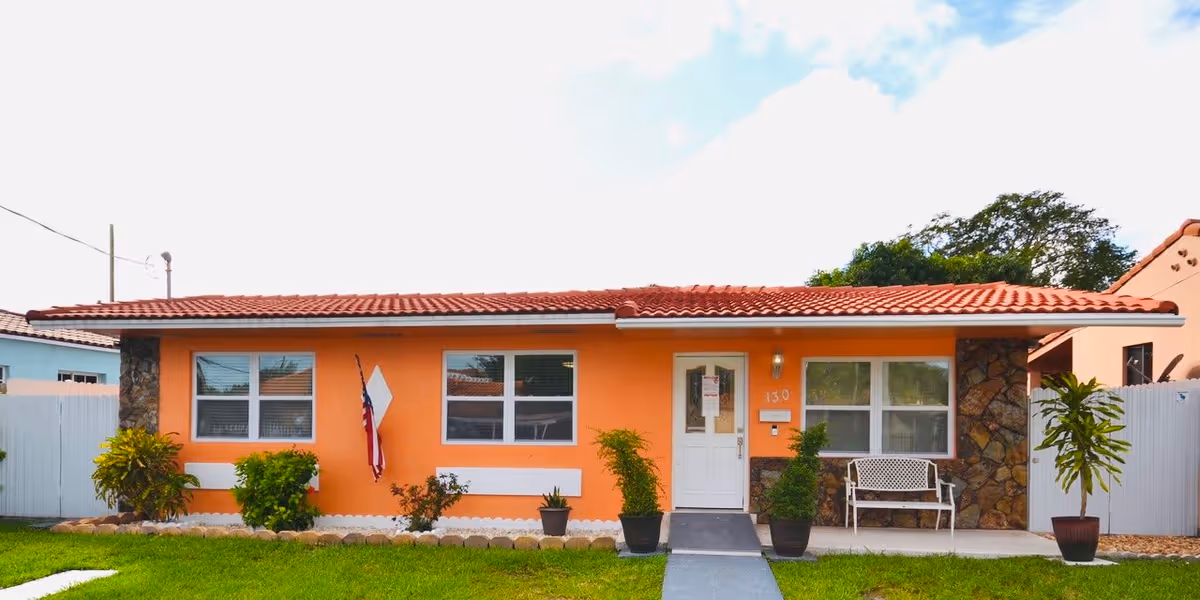 Single-story orange building with a red tile roof, white door, and three windows. There are several potted plants and bushes in front of the building, a white bench near the entrance, and an American flag hanging on the wall. The building is surrounded by a white fence and has a green lawn in front.