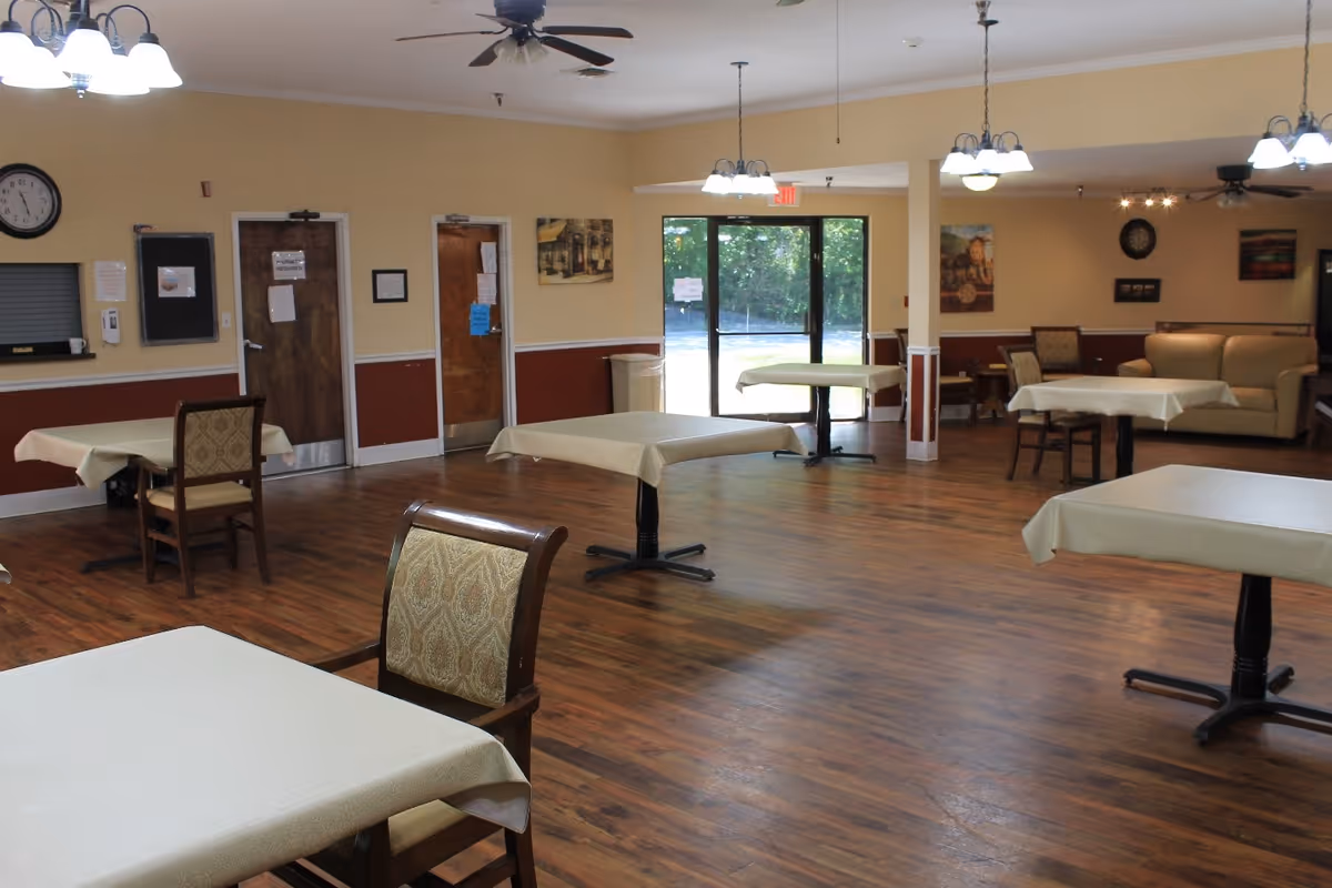 A spacious dining room with several tables covered with beige tablecloths and wooden chairs with patterned cushions. The room has hardwood flooring, ceiling fans, and multiple light fixtures. There is a glass door leading outside and a few framed pictures on the walls.