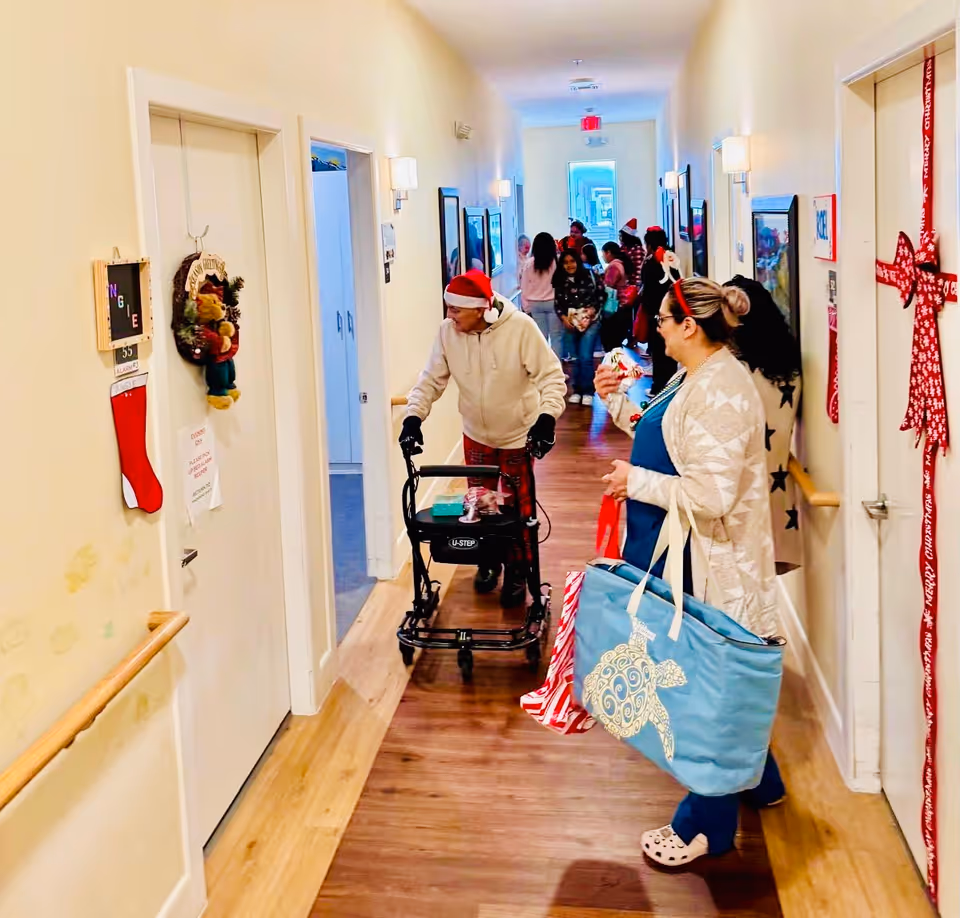 A decorated hallway in an assisted living facility with holiday decorations on the doors and walls. Several people, including an elderly person using a walker and a woman carrying bags, are present. The hallway has wooden floors and light-colored walls with handrails.