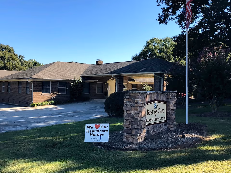 Brick assisted living building with a stone sign reading 'Best of Care Assisted Living Center', a small lawn sign honoring healthcare workers, and an American flag.