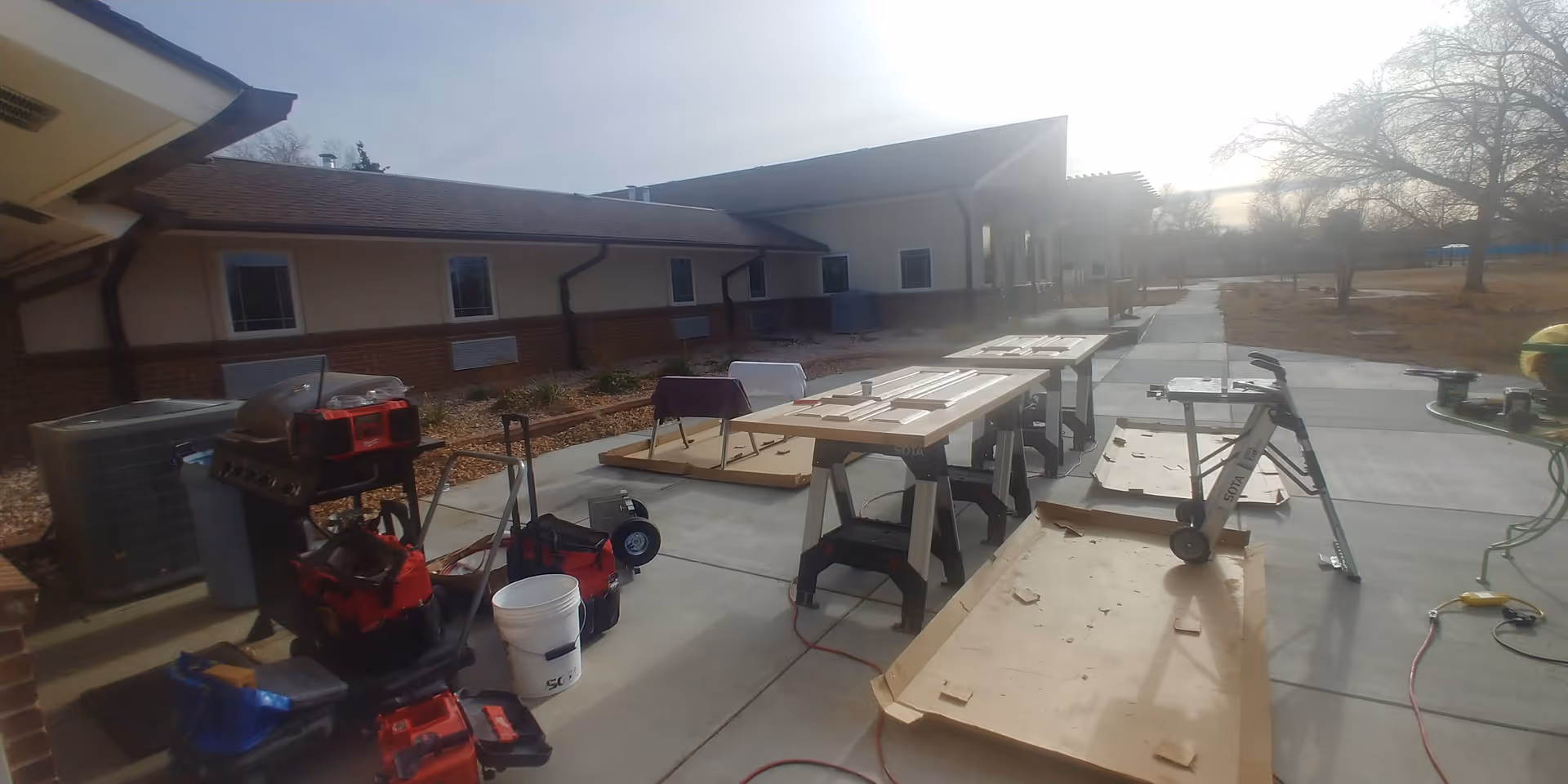 Outdoor patio area of a facility with construction or maintenance equipment and materials set up on the concrete walkway. Several tables hold wooden doors or panels, and various tools, buckets, and carts are scattered around. The building exterior is visible with windows and a covered walkway extending into the distance. Trees and open land are in the background under a bright sky.
