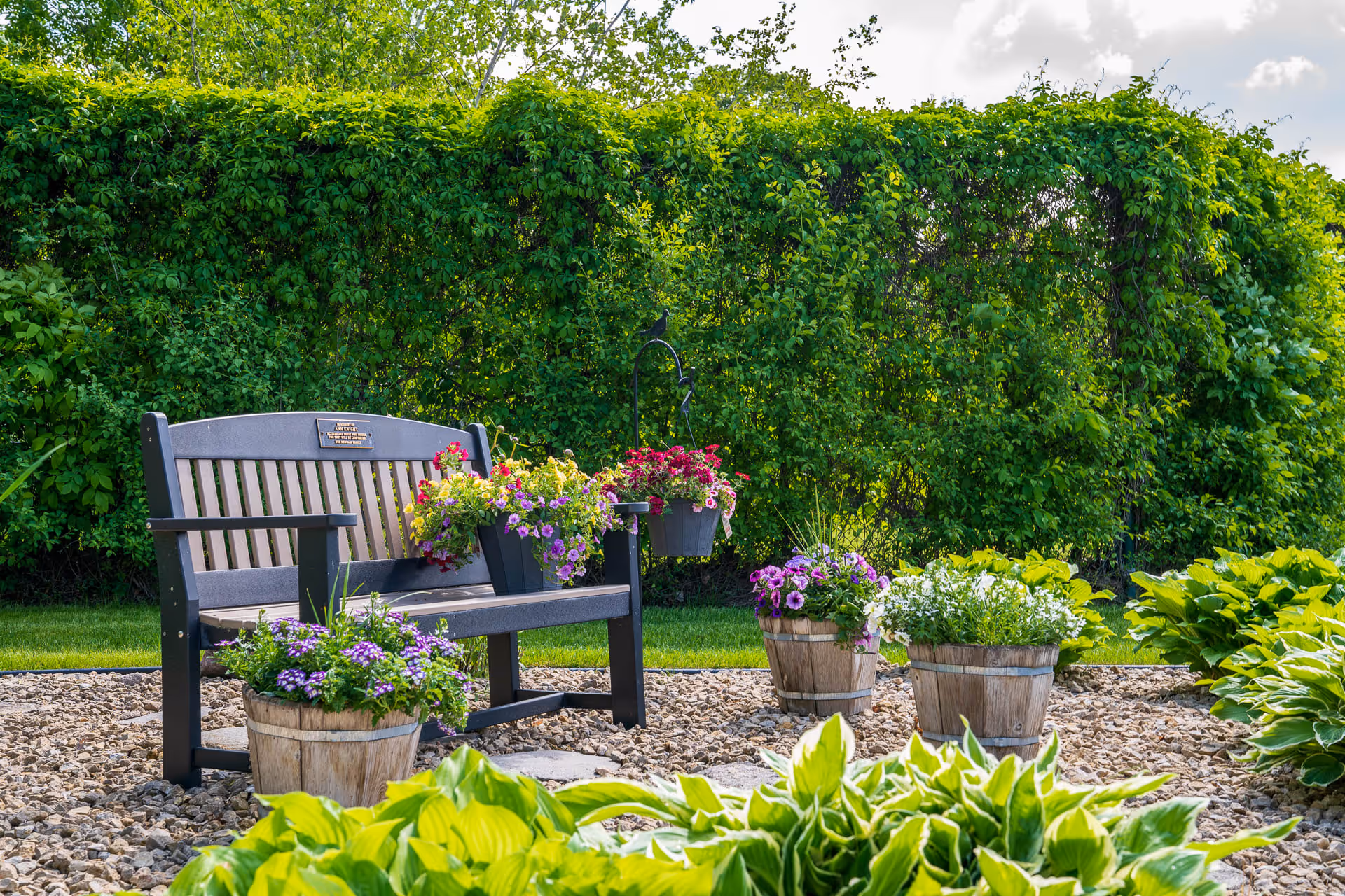 A black wooden bench with a memorial plaque is placed in a garden area surrounded by green bushes and plants. The bench is decorated with colorful flower pots containing purple, pink, yellow, and white flowers. The ground is covered with small stones and stepping stones, and the background shows a clear sky with some clouds.