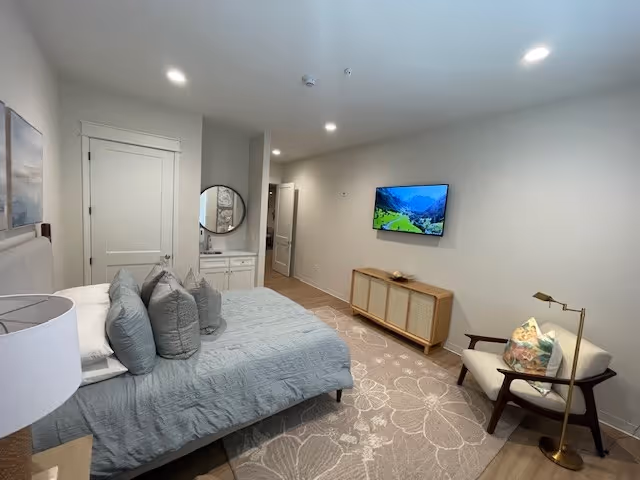 A modern bedroom with a neatly made bed featuring multiple gray pillows and a gray bedspread. There is a round mirror above a small white cabinet near the door. A flat-screen TV is mounted on the wall opposite the bed. A light-colored armchair with a decorative pillow and a gold floor lamp are positioned near the TV. The room has light-colored walls, recessed ceiling lights, and a large floral-patterned area rug on the wooden floor.