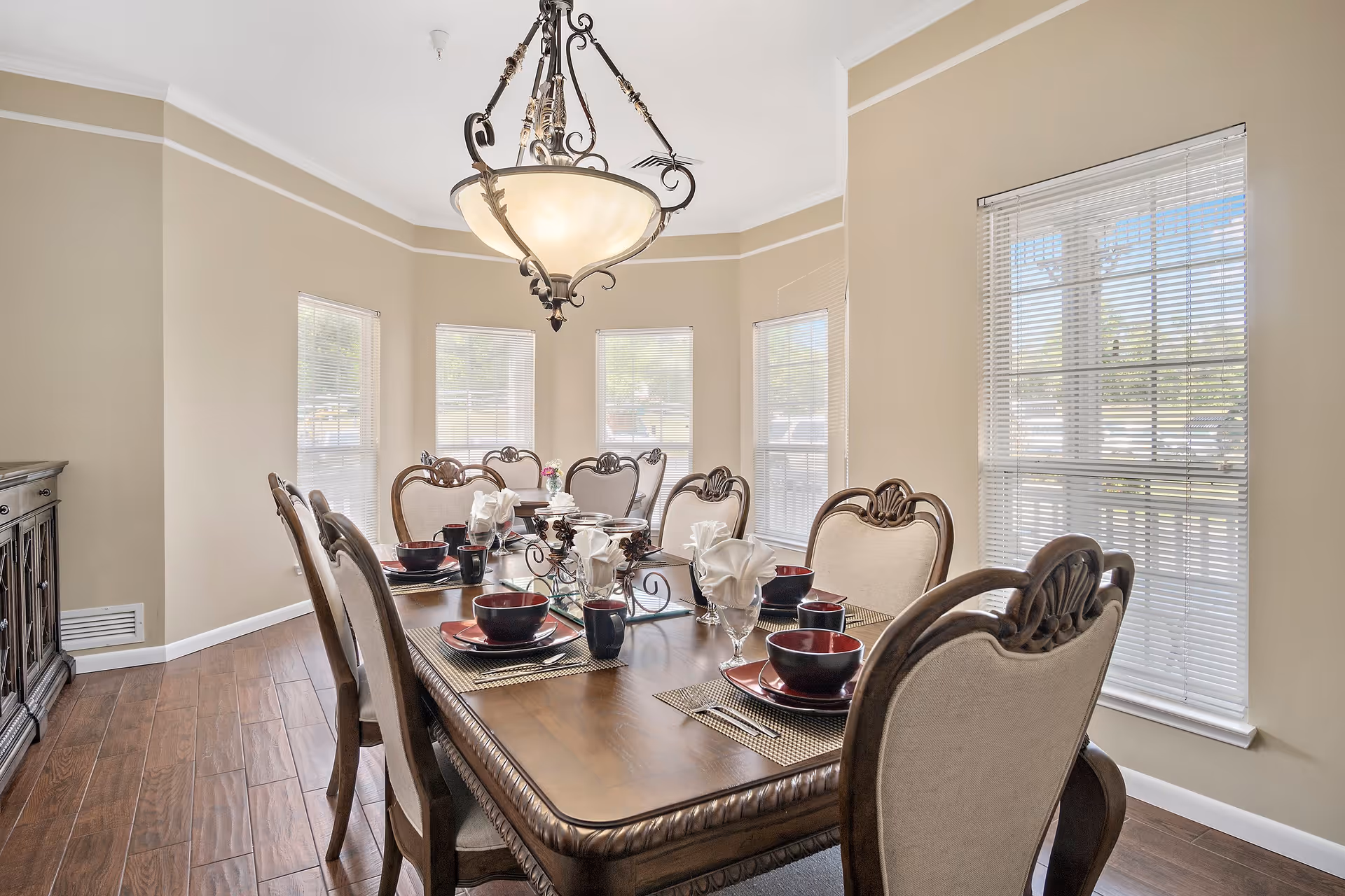 A well-lit dining room with a large wooden dining table set for six people. The table features elegant place settings with black and red bowls, plates, cups, and folded white napkins. The room has beige walls, multiple windows with blinds allowing natural light, a wooden sideboard, and a decorative chandelier hanging above the table.