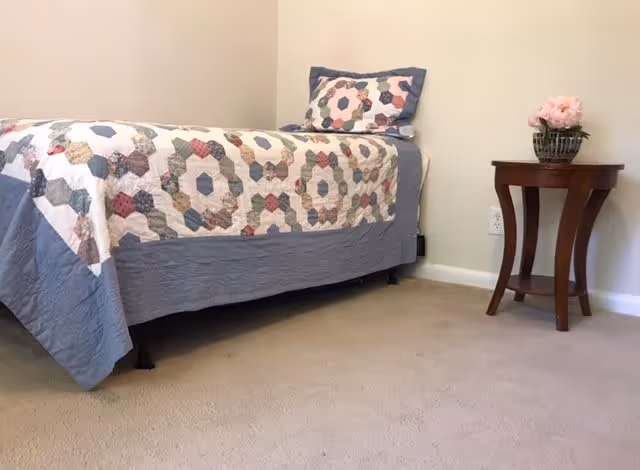 A simple bedroom with a single bed covered in a patchwork quilt with hexagonal patterns in various colors. There is a matching pillow on the bed. Next to the bed is a small wooden side table with curved legs, holding a basket with pink flowers. The walls are plain and light-colored, and the floor is carpeted.