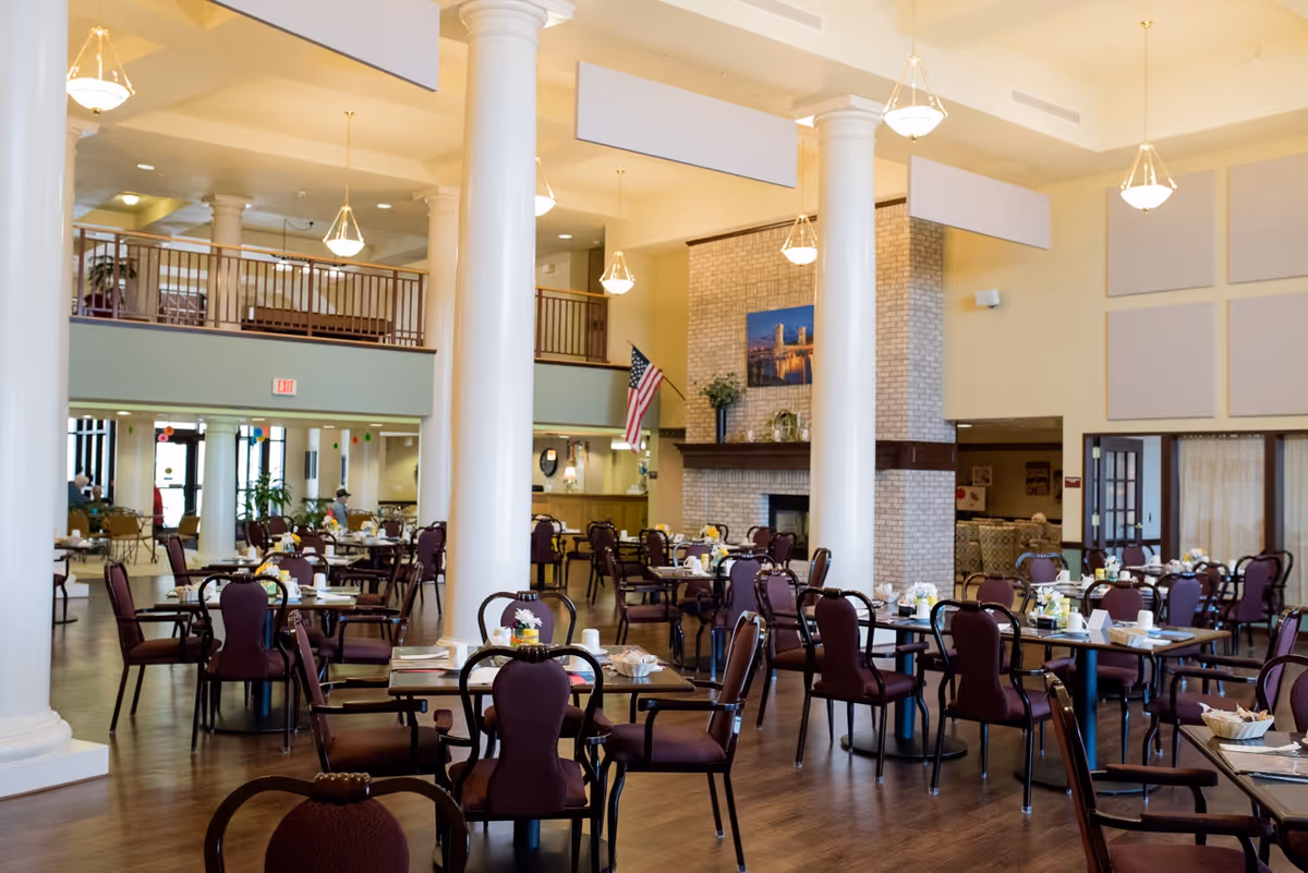 Large dining room with many set tables and burgundy chairs, tall white columns, and a fireplace.