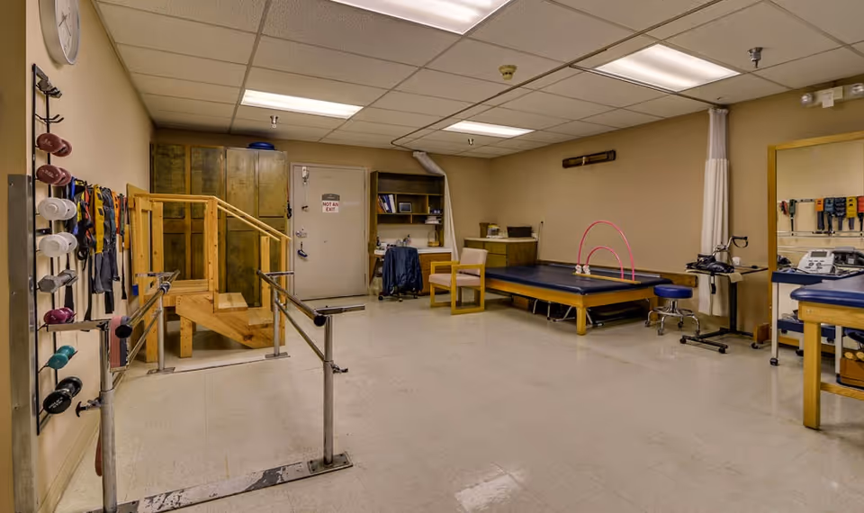 A physical therapy room with parallel bars for walking exercises, a small wooden staircase, therapy mats, chairs, and various exercise equipment mounted on the walls. The room has beige walls, a tiled floor, and fluorescent ceiling lights.