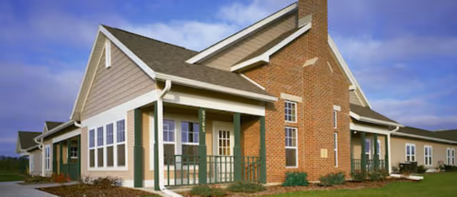 Exterior view of a single-story senior living facility building with beige siding and brick accents, featuring multiple windows and a covered porch area under a partly cloudy sky.