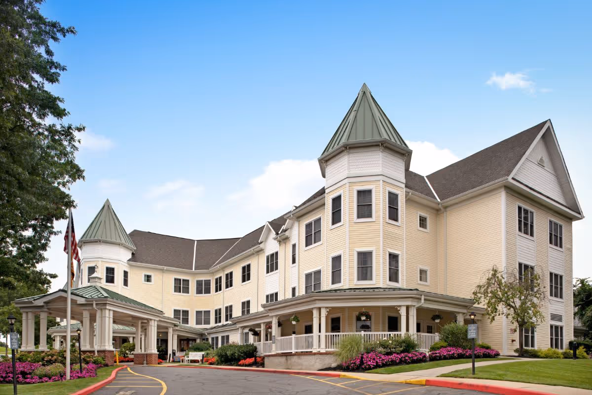 Front exterior of a multi-story light-yellow senior living building with turrets, a covered porte-cochere entrance, and landscaped flowerbeds.