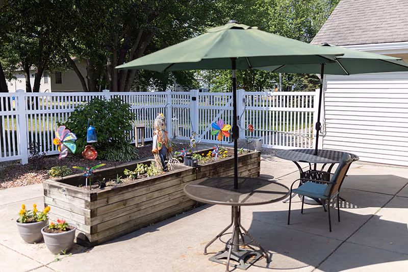 Outdoor patio area with two round metal tables, each shaded by a green umbrella. One table has a chair with a cushion. There is a raised wooden planter box with colorful pinwheels, flowers, and a small scarecrow decoration. The area is enclosed by a white fence and surrounded by trees and greenery.
