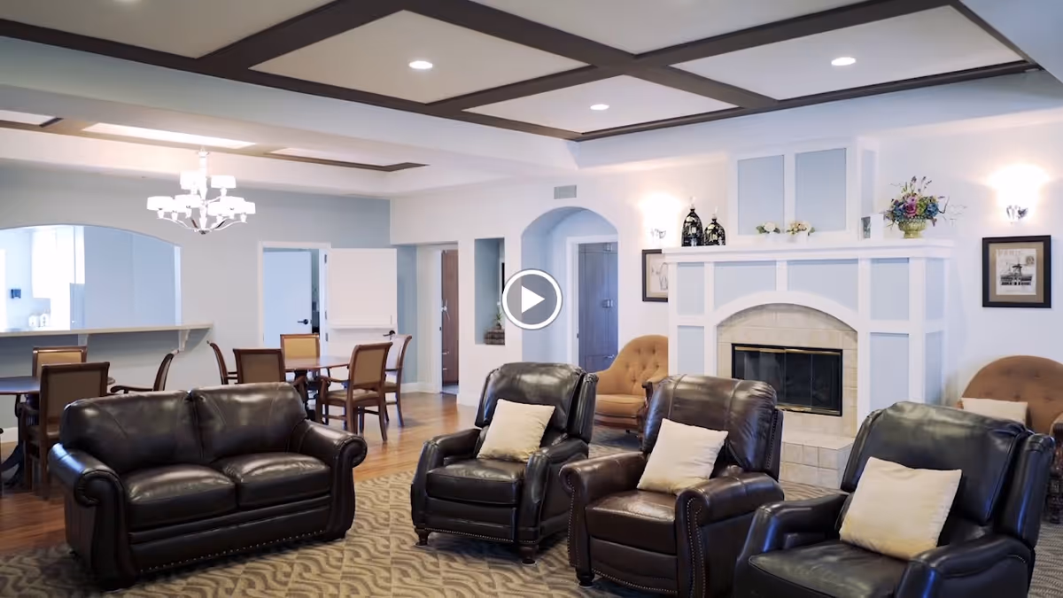 A spacious and well-lit living room area with dark leather sofas and armchairs arranged around a light-colored fireplace. The room features a coffered ceiling with dark wood beams, a chandelier, wooden flooring, and a dining table with chairs in the background. The walls are painted light blue and white, decorated with framed pictures and floral arrangements.