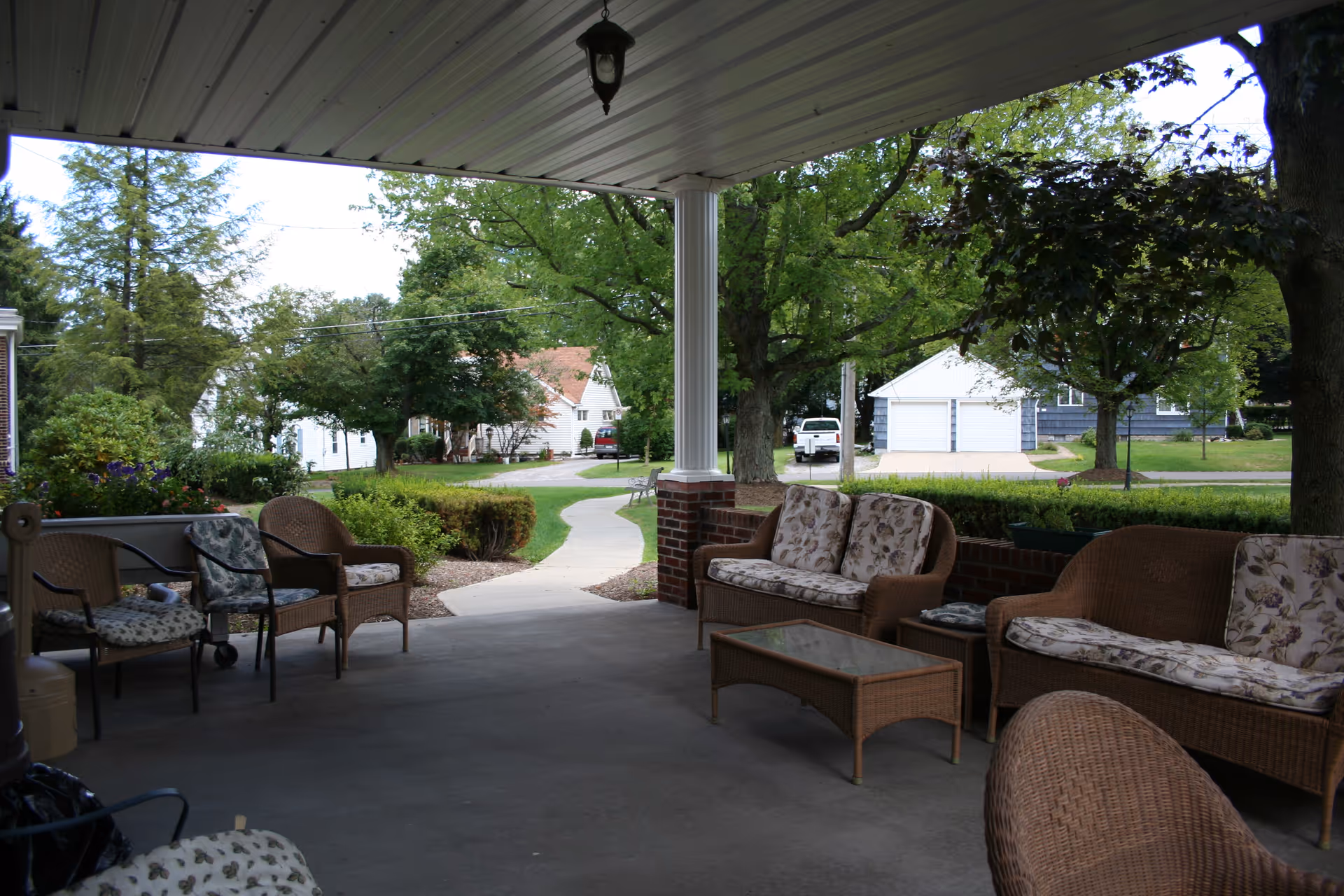 Covered outdoor patio area with wicker furniture including chairs and sofas with floral cushions, overlooking a walkway and green trees in a residential neighborhood.