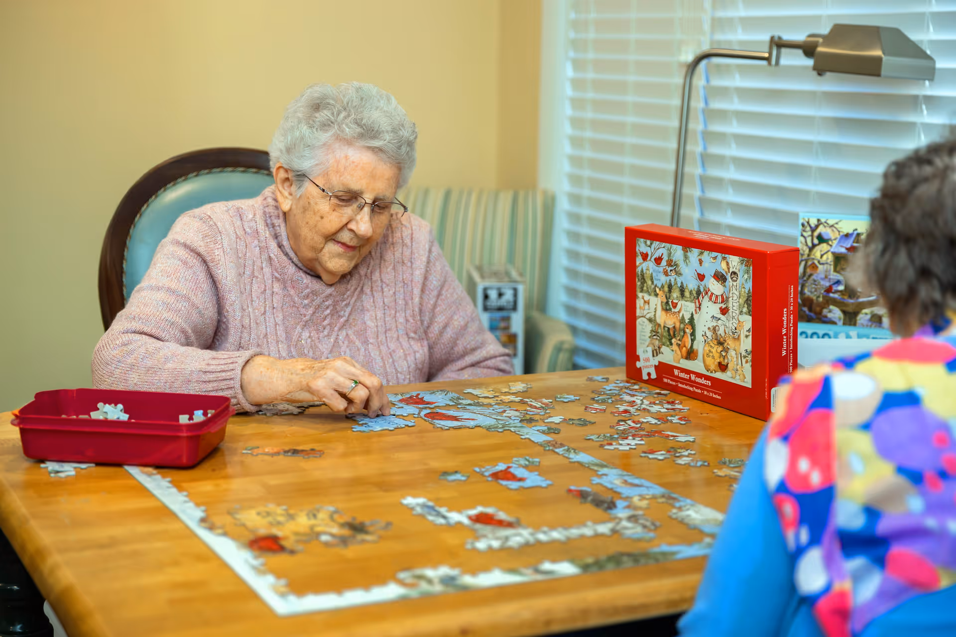 An elderly woman with gray hair and glasses is sitting at a wooden table working on a jigsaw puzzle. She is wearing a pink sweater and is focused on placing puzzle pieces. Another person, partially visible from behind, is sitting across the table wearing a colorful patterned top. A puzzle box with a winter-themed image is on the table near a window with white blinds. A lamp is positioned above the puzzle box.