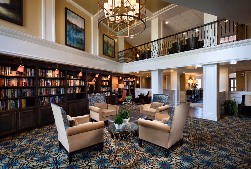 Two-story library-style lounge with built-in bookshelves, a seating area of armchairs around a glass table, a large chandelier, and an upper balcony railing.