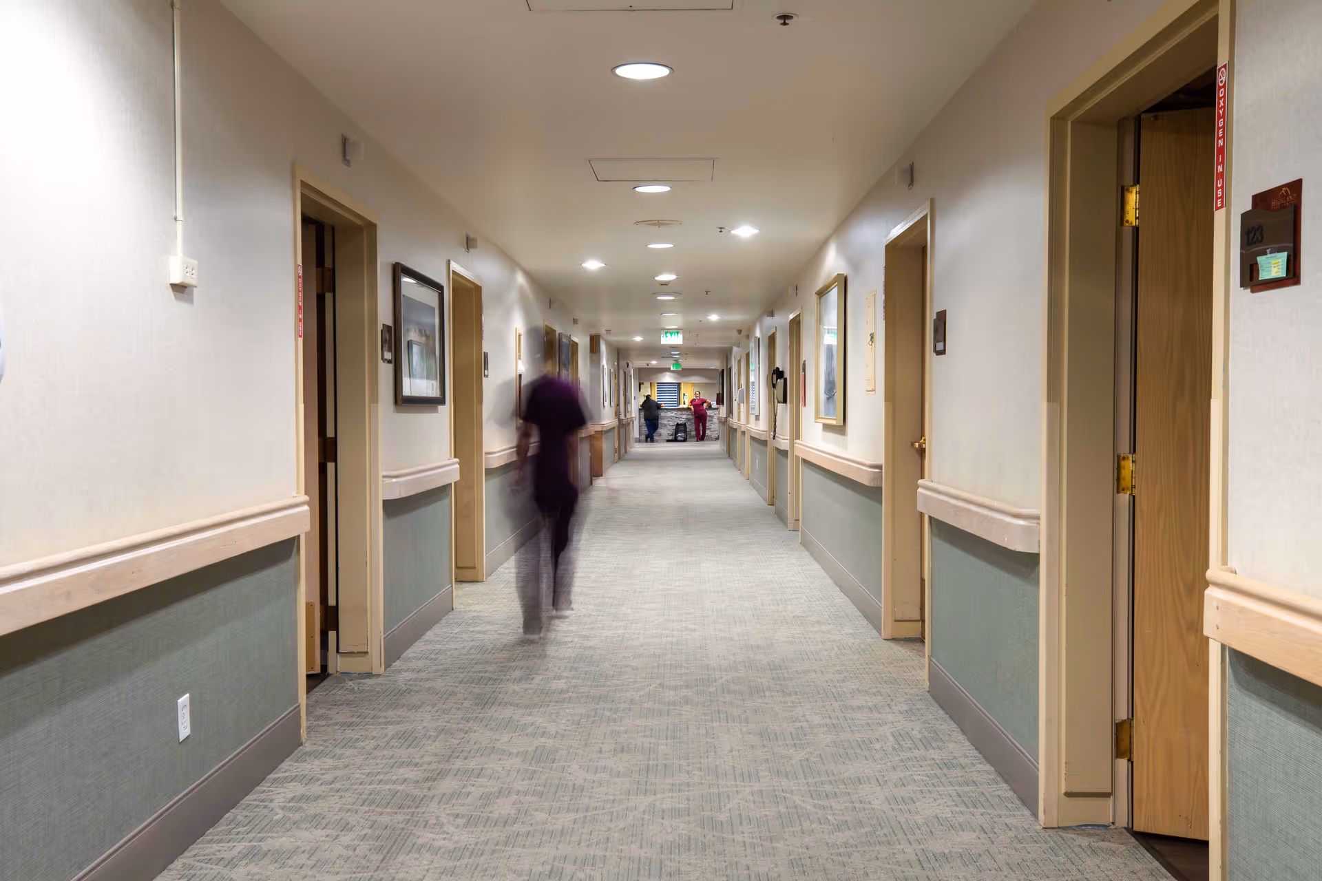 A long, well-lit hallway in a care facility with multiple doors on both sides, handrails along the walls, framed pictures, and a few blurred figures walking or standing in the distance.