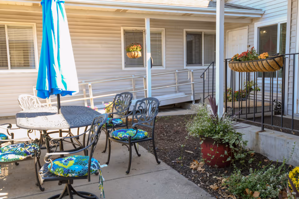 Outdoor patio area with a metal table and chairs featuring colorful blue and green cushions. A large blue umbrella is attached to the table. The patio is adjacent to a building with beige siding, windows, a ramp, and a door. There are potted plants and hanging flower baskets adding greenery and color to the space.