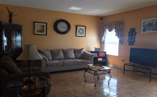 Sunlit living room with beige sofas, a wall clock and framed art, a coffee table, TV on a stand, and tiled floor.