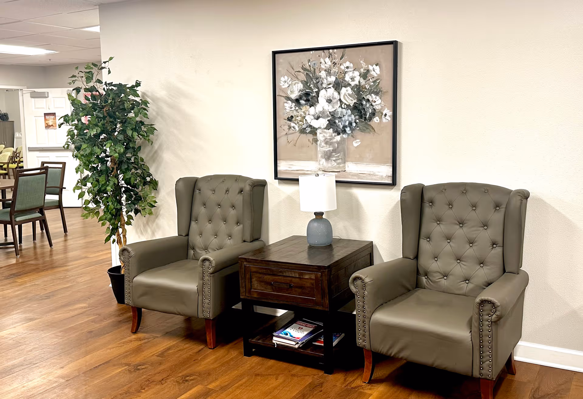 Two olive green tufted armchairs with nailhead trim are placed on either side of a dark wooden side table with a drawer and a blue lamp on top. Behind the chairs is a large framed painting of white flowers in a vase. To the left, there is a tall green potted plant and a glimpse of a dining area with wooden chairs and tables. The floor is wooden and the walls are light-colored.