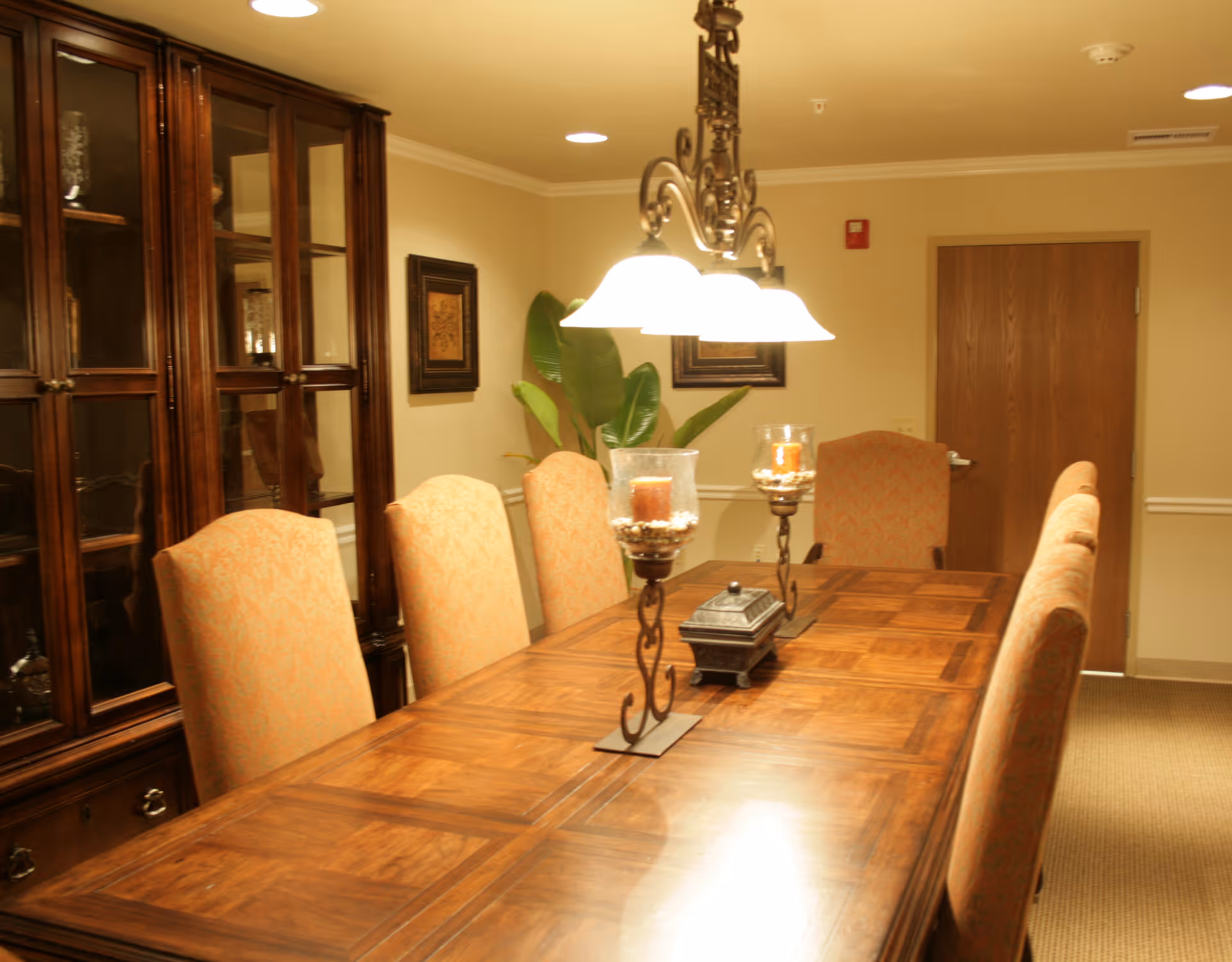 A warmly lit dining room with a long wooden table surrounded by upholstered chairs. The table has decorative candle holders and a small box. A wooden cabinet with glass doors is visible on the left side, and framed artwork and a large green plant are on the wall in the background. A closed wooden door is at the far end of the room.
