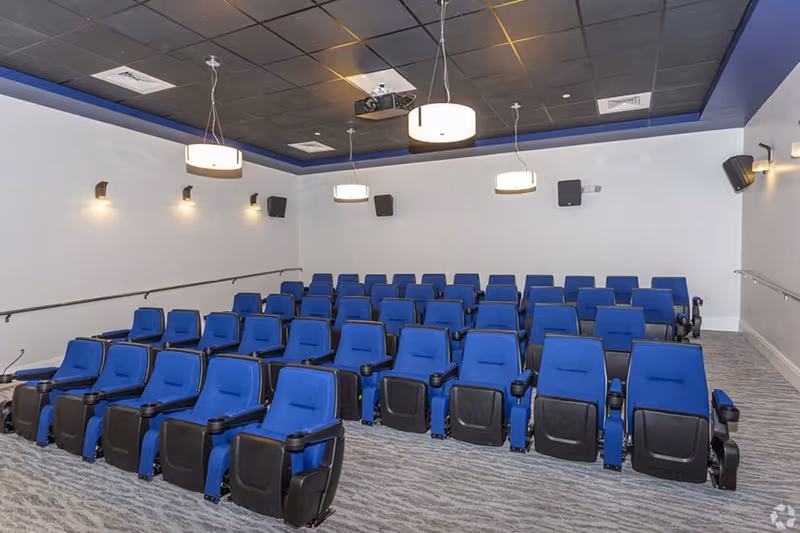 A small theater room with four rows of blue cushioned seats arranged in a tiered layout. The room has a dark ceiling with three hanging circular light fixtures and wall-mounted speakers. The walls are white with small wall lights and handrails on both sides.