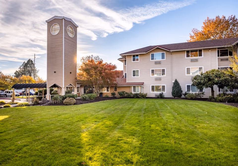 Three-story retirement facility building with a tower featuring round windows, a well-kept lawn and landscaped grounds under a blue sky.